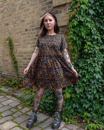 A person with tattoos holds out their Run & Fly oversized smock dress, showcasing its flowing fabric with a spooky Halloween design. The print features orange and green leaves, skulls, and stars. The person wears fishnet tights and platform boots. The brand is a small ethical slow fashion company.