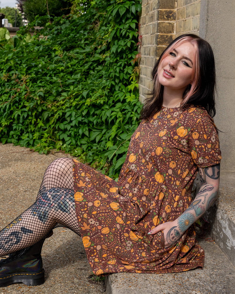 A person sits outdoors in a Run & Fly smock dress with a brown fabric covered in pumpkins, mushrooms, and leaves. Their arms and legs feature tattoos, and they are wearing fishnet tights and boots. The setting includes a brick wall and green foliage. The brand is a small ethical slow fashion company.