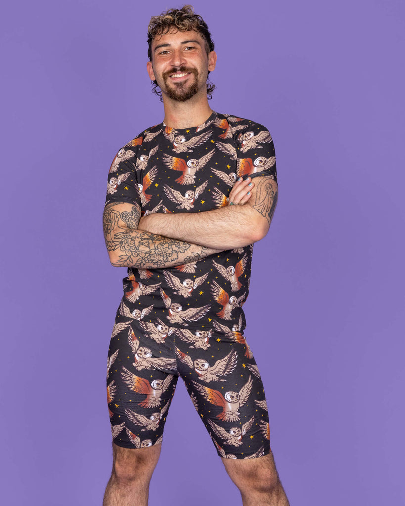 The What a Hoot Owl Stretch Cycle Shorts worn by a non binary masc person with a short curly brown mullet wearing the matching tshirt and white trainers on a purple studio background. They are facing forward smiling with their arms crossed in front of them. The black base shorts features brown tawny owls flying amongst yellow stars.