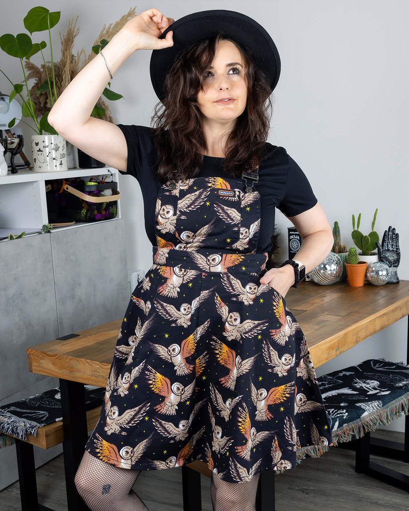 Amy, a femme model brunette hair, is in a studio in Hove wearing What A Hoot Owl Flared Pinafore Dress with a black t shirt underneath, a black fedora hat and black fishnet tights with chunky boots. The black pinafore has an all over print of cute barn owls and yellow stars. Amy is leaning against a table with one leg bent and has one hand in the dress pocket and other on the brim of her hat.