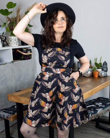 Amy, a femme model brunette hair, is in a studio in Hove wearing What A Hoot Owl Flared Pinafore Dress with a black t shirt underneath, a black fedora hat and black fishnet tights with chunky boots. The black pinafore has an all over print of cute barn owls and yellow stars. Amy is leaning against a table with one leg bent and has one hand in the dress pocket and other on the brim of her hat.