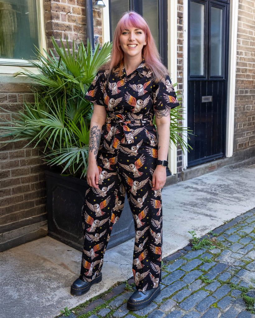 Model Stacie poses outside in a black jumpsuit with a bold owl and stars print. She stands confidently with pink hair, smiling, against a brick and cobbled background with greenery.