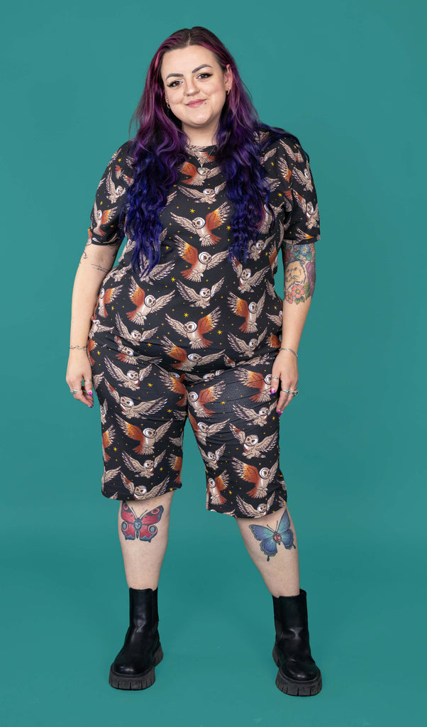 The What a Hoot Owl Stretch Cycle Shorts worn by a femme alternative tattooed model with long pink and purple hair, the matching tshirt and black boots on a teal studio background. She is facing forward smiling with both hands resting by her sides. The black base shorts features brown tawny owls flying amongst yellow stars.