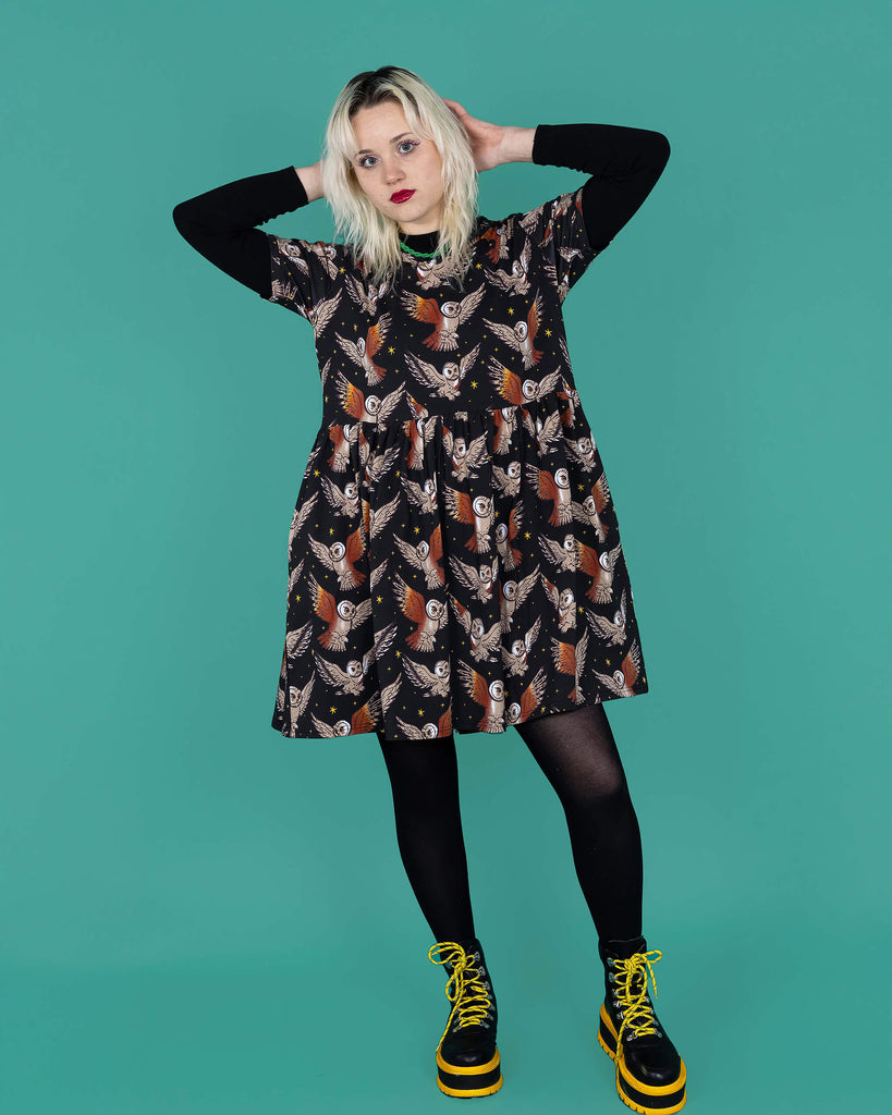 The What a Hoot Owl Oversized Smock Dress worn by a femme model with white blonde hair, a long black tshirt, black tights and yellow black boots on a teal studio background. She is facing forward with both hands resting behind her head with one leg bent forward and smising to camera. The black base dress features brown tawny owls flying amongst yellow stars.