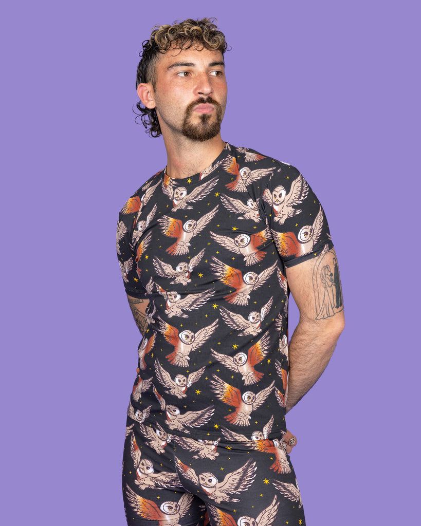 The What a Hoot Owl Short Sleeve T Shirt on a non binary masc presenting person with a short brown curly mullet haircut and the matching cycle shorts on a purple studio background. They are facing forward pouting looking off to the right with their arms crossed behind their back. The black base tshirt features brown tawny owls flying amongst yellow stars.