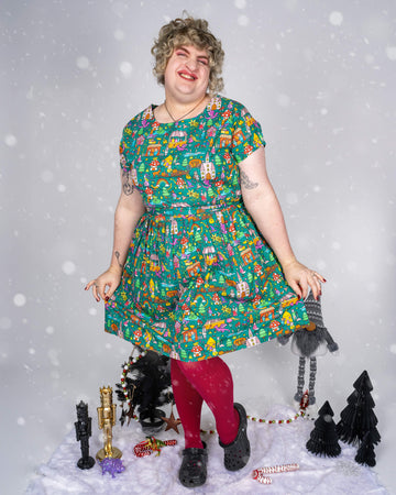 Model Maddie stands against a soft, snowy-themed background, smiling brightly while wearing a vibrant green dress adorned with a whimsical fairytale scene print by Waffle Mama Design. The dress features various colourful characters, animals, and storybook houses. Maddie pairs the dress with bold red tights and black shoes, radiating a cheerful and festive energy.