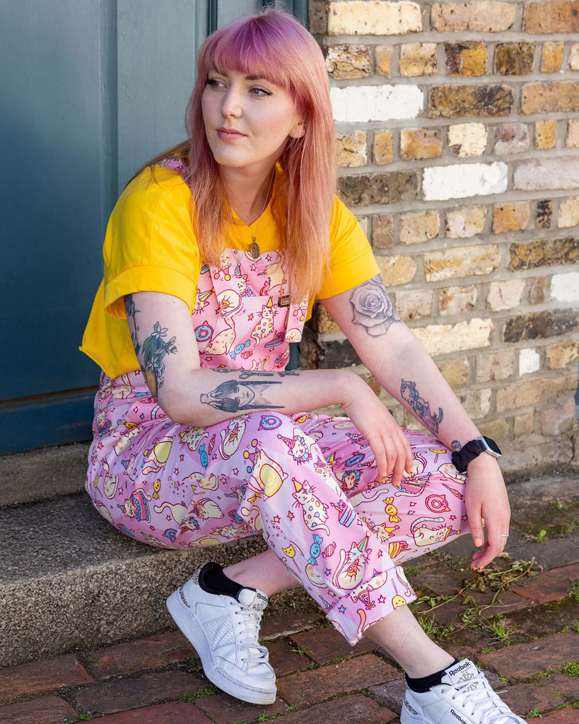 Stacie is in a mews in Hove wearing Run & Fly x The Mushroom Babes Party Cats Stretch Twill Dungarees with a bright yellow top underneath and white trainers. The dungarees are baby pink with white cats with party hats on and eating birthday cake, as well as various pink, blue and yellow sweets designed by The Mushroom Babes. Stacie is sat on a step with her ankles crossed and arms resting on her legs. She is looking off to one side.