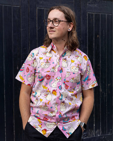 Michael a man with shoulder length hair and glasses is looking off to one side wearing a pink shirt with party cats on against a black door.