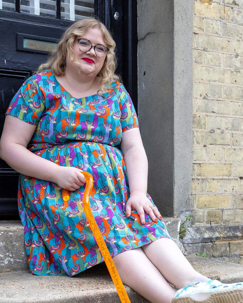 Model is wearing the Run & Fly Party Llama stretch belted tea dress, with waist belt tie, side pockets and zip, and blue, green, orange and purple Llama print pattern. The model is sat on steps outside a town house in Hove smiling with their ankles cross and holding their orange walking aid. 