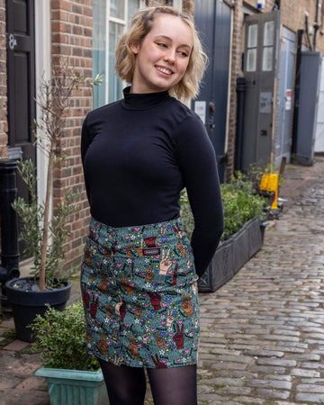 Model with short blonde hair wearing Peace and Love A Line Skirt paired with long sleeve black turtleneck top and chunky black boots. The skirt an olive green colour with hands in different skin tones making the peace sign, various flowers, peace symbols and love hearts in white, yellow, purple and pink and 'peace' and 'love' printed on. There is a Run&Fly logo on front left pocket of the skirt and it has silver buttons down the front.