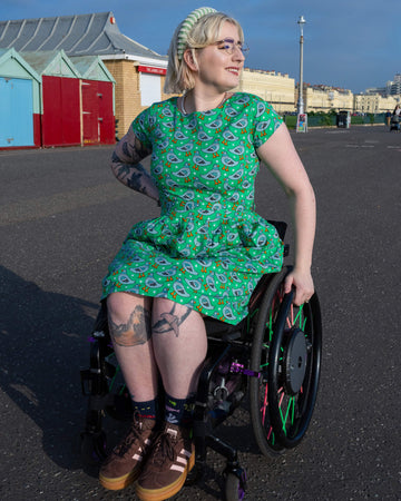 Eliza is sat outside in their wheelchair by Hove promenade wearing Run & Fly Pigeon Stars belted tea dress with pockets  with brown trainers. The bright green dress has an all over print of pigeons and white stars. They are posing with one hand on their hip and the other on the wheel of the wheelchair. They are smiling looking off to one side