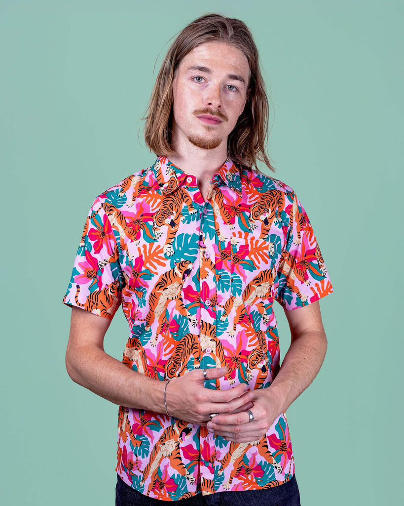 Jack is stood in a photography studio in Hove with a green backdrop wearing Tiger Lily Stretch Short Sleeve Shirt with jeans. The baby pink shirt has an all over print of tigers with flowers and leaves. Jack is posing toward the camera with his hands together.