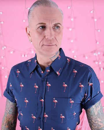 Jim is stood in front of a pink studio background wearing the preppy flamingo short sleeve shirt with burgundy trousers. They are heavily tattooed and have swept back silver hair. They are facing the camera posing with both arms out by their side and looking off to the right. Photo is cropped from the hips up.