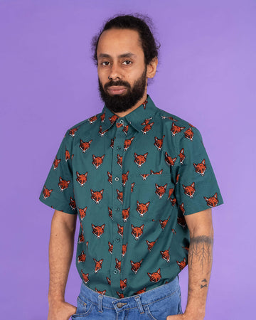 Richard, a hispanic male model with dark hair in a bun and a beard, is stood in a photography studio in Hove in front of a lilac backdrop wearing Preppy Foxy Short Sleeve Shirt with blue jeans. The emerald green shirt has an all over print of fox heads. Richard is posing facing the camera with his thumbs resting in the front pockets of his jeans. The photo is cropped at the thighs.