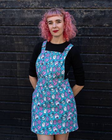 A woman with pink curly hair stands confidently in a turquoise "Pretty Spooky" pinafore dress by Run & Fly. The dress is covered in pastel ghosts, crystals, and skull motifs, set against a black wooden background. She pairs the look with a black long-sleeve top, showcasing Run & Fly's ethical slow fashion ethos.