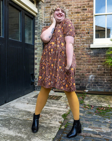 Maddie stands outdoors, wearing the Run & Fly pumpkin dress, holding a matching tote bag with a similar autumnal design. The dress has short sleeves and a relaxed fit, perfect for a cosy autumn look.