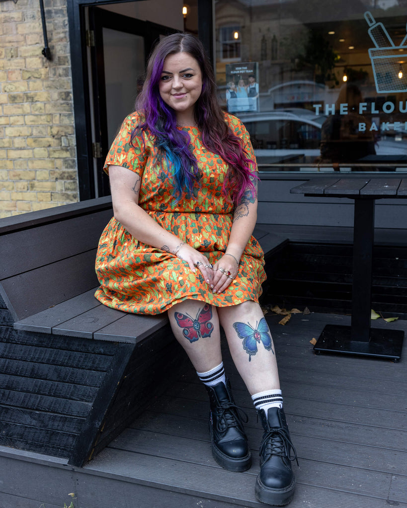 Luisa, a white femme model with colourful hair, is wearing Orange Cactus Tea Stretch Dress with Pockets paired with chunky black boots. The background colour of the dress is a bright orange with various cactus patterns on it in different shades of green. Luisa is sat on a bench outside a bakery in Hove with her ankles crossed and is smiling at the camera. 