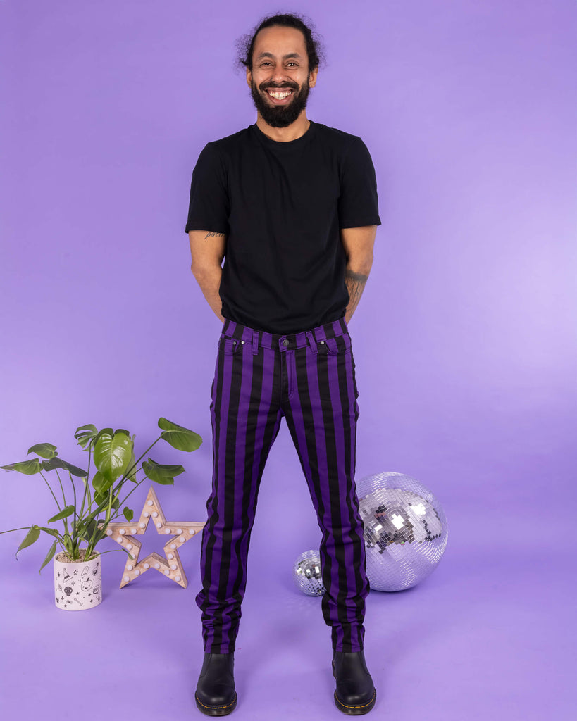  Model Richard stands against a purple backdrop, smiling warmly while wearing Run & Fly’s bold purple and black striped trousers. He pairs the statement trousers with a simple black T-shirt and black boots. The background includes a plant, a decorative star light, and disco balls, adding a playful and vibrant touch to the scene.