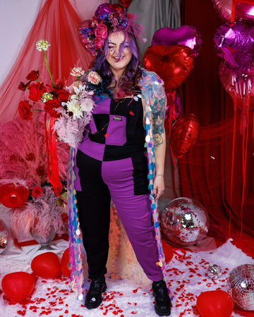 Luisa, a white femme model with purple, blue and pink in their hair is stood in a studio in Hove with a red backdrop with heart shaped balloons and pink feathers and rose petals on the floor. They are wearing Purple and Black Mix n Match Two Tone Stretch Twill Dungarees with a sequin shawl and floral headdress. Luisa is posing holding flowers and facing the camera. 