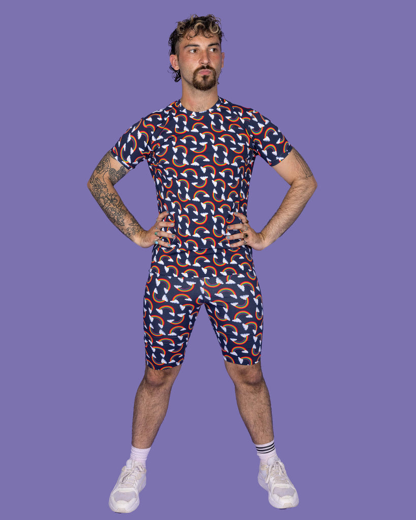 The Over the Rainbow & Clouds Short Sleeve T Shirt worn by a masc non binary person with a brown curly mullet, the matching cycle shorts and white trainers on a purple studio background. They are facing forward pouting looking off to the right with both hands on their hips. The navy blue base tshirt has repeating rainbow arches with white clouds.