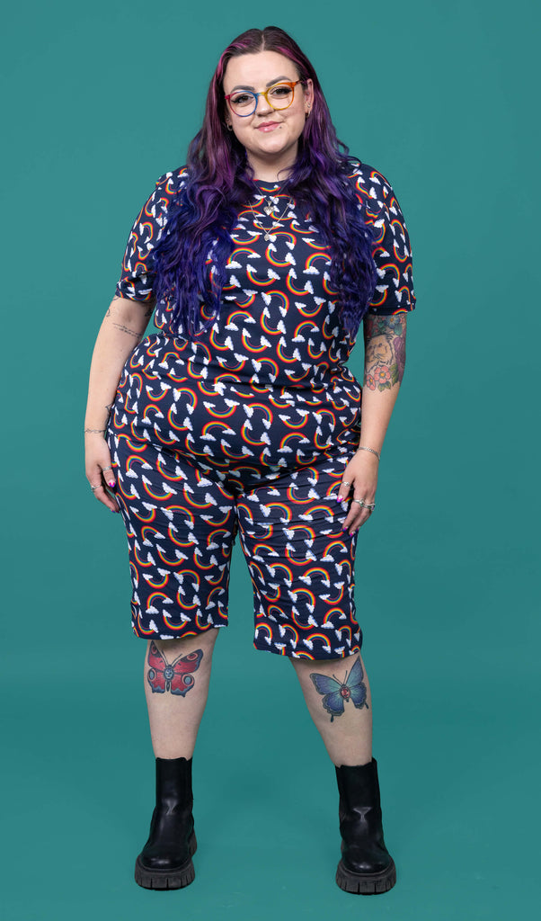 The Over the Rainbow & Clouds Stretch Cycle Shorts worn by a femme alternative tattooed model with rainbow glasses, long pink to purple hair, the matching tshirt and black boots on a teal studio background. She is facing forward smiling with both arms resting by her side. The navy blue base shorts has repeating rainbow arches with white clouds.