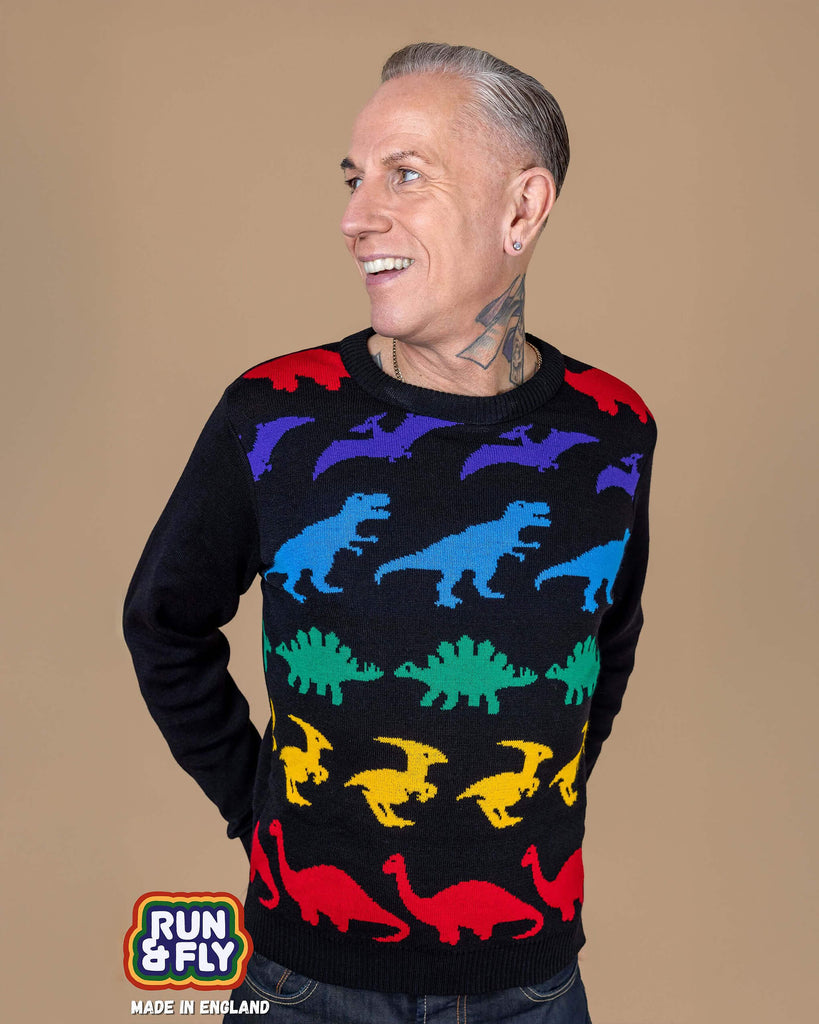 James is wearing Rainbow Dinosaur Mash Up Jumper with jeans. The jumper is a plain black base with various dinosaurs in a rainbow colour stripe sequence with plain black arms and back. James is stood in front of a beige backdrop posing with his hands behind him and smiling looking off to one side. 