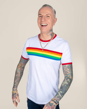 James smiling warmly, wearing a white T-shirt with a bold rainbow stripe across the chest and red trim on the sleeves and collar, paired with dark jeans and accessorised with gold jewellery.