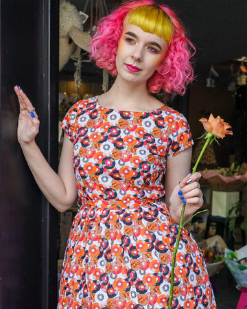 Lottie is stood in front of a flower shop holding a peach rose wearing the vinyl record print off white tea party dress. She is facing forward posing leaning against a black wall and looking off to the left smiling. Photo is cropped from the knees up.