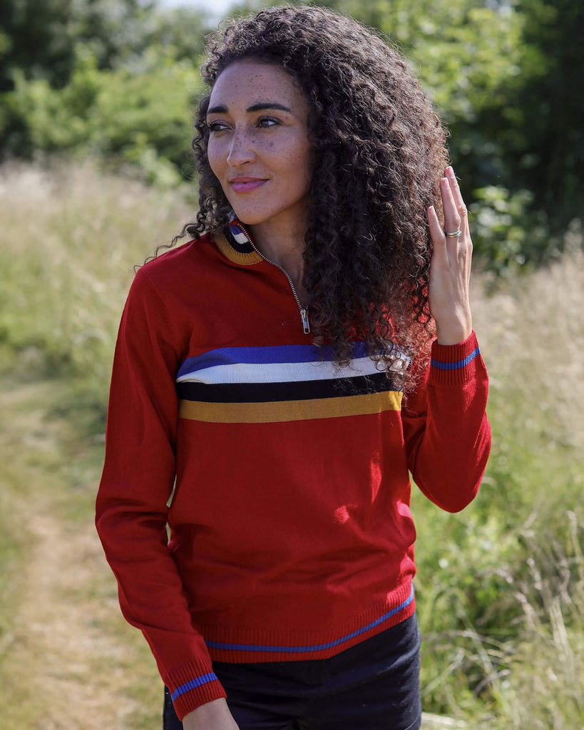 Rochelle is stood in a grass field wearing the red retro zip neck jumper with black shorts. They are facing forward posing with one hand running through their hair and the other by their side whilst smiling to the left. Photo is cropped from the hips up.