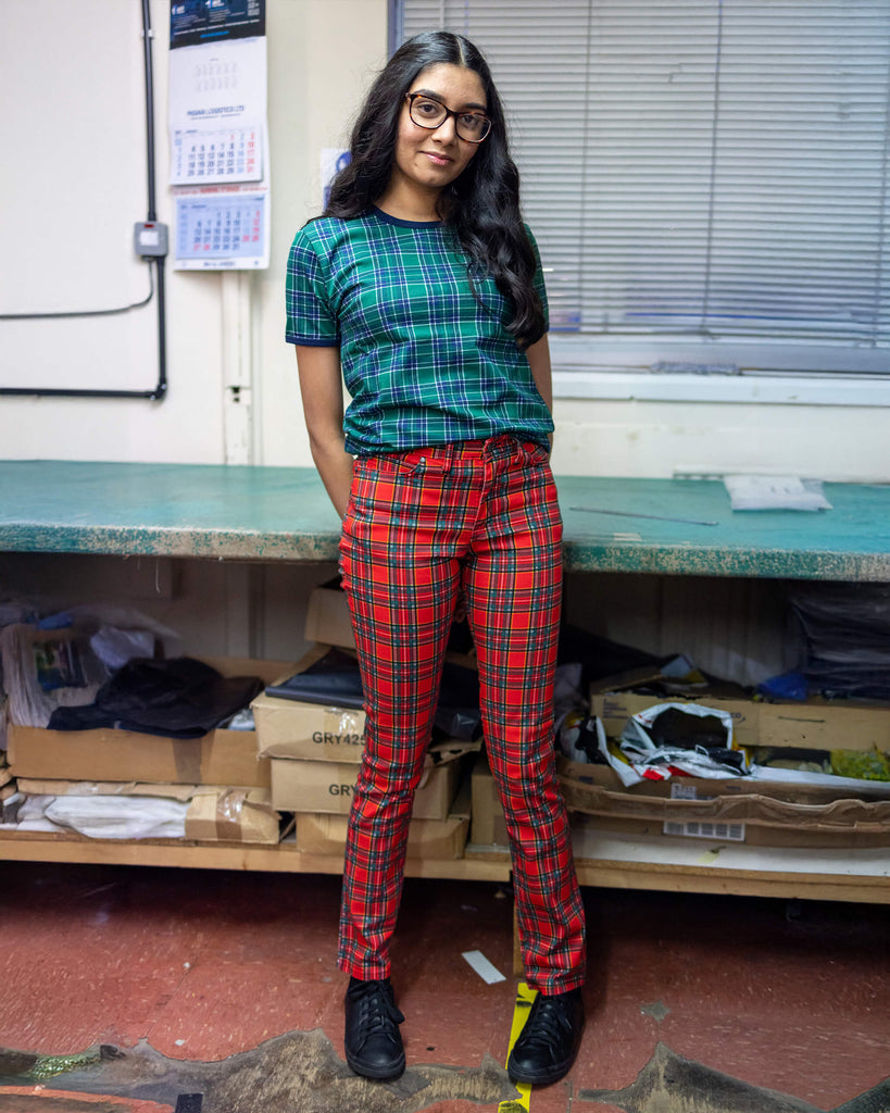 Model is stood in our warehouse area wearing the mid rise red tartan stretch skinny jeans with a green tartan tshirt and black trainers. They are facing towards the camera leaning against a desk with both hands behind their back and smiling.