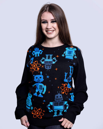 Charlotte is stood in front of a grey sofa wearing the robot jumper with black stone wash flares. She is facing forward with both hands resting by her side and smiling to camera. Photo is cropped from the thighs up. The robot jumper is a black base featuring multiple blue robots with orange, blue stars and chemical symbols with plain black arms and back.