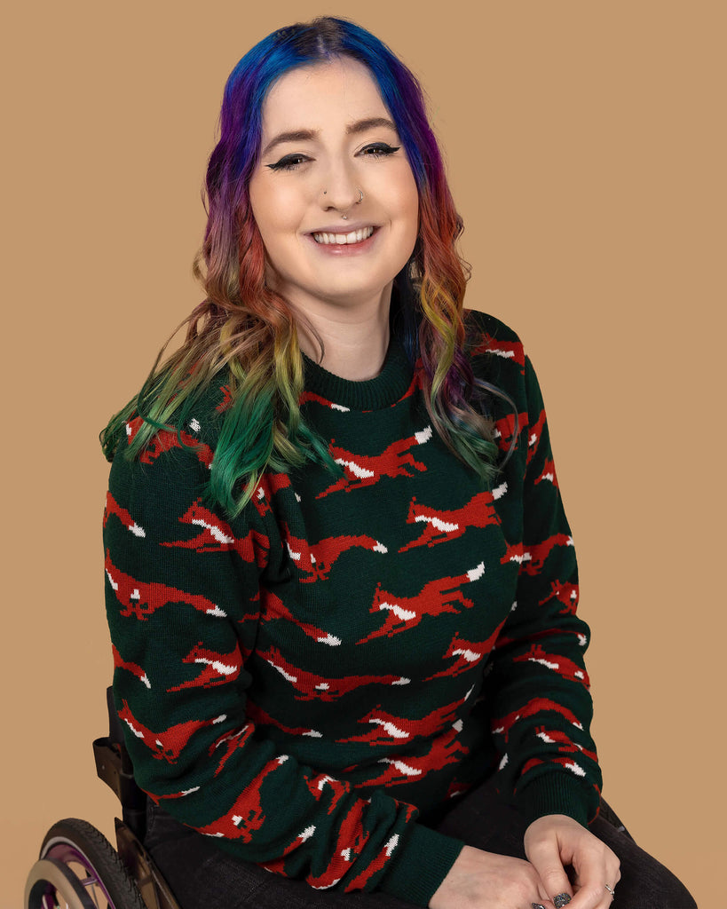 Eliza, a femme model with rainbow coloured hair, is sat in their wheelchair in front of a tan backdrop with flowers on it. They are wearing the Running Fox Jumper and smiling at the camera. 