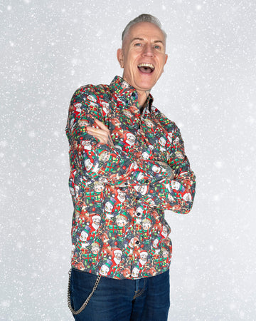 A cheerful model wearing a Run & Fly Christmas shirt with a retro holiday print stands with arms crossed against a snowy background. The quirky print features Santa, snowmen, elves, and festive decorations, making it a playful ethical slow fashion choice for the holidays.