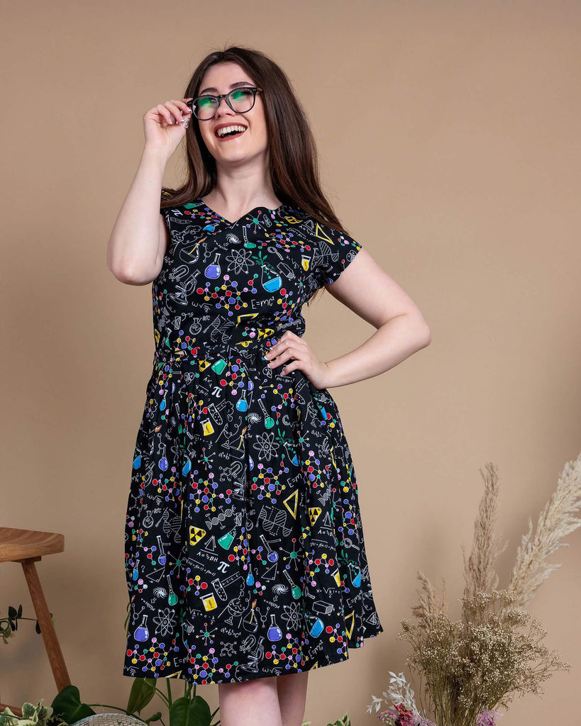 Charlotte is stood in front of a beige backdrop with various plants and disco balls behind her. She is wearing School of Science Belted Tea Dress with Pockets with black boots. The dress has a black background colour with an all over print of various chemistry equipment in white, yellow, green, red, yellow and purple. Charlotte is smiling and holding onto her glasses with her ankles crossed and hand on her hip.