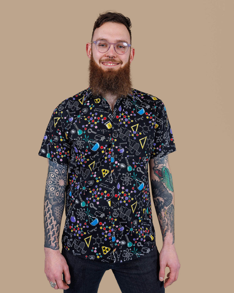 Dec is wearing the science print short sleeve shirt, featuring multicoloured science symbols on black. Model is posing in studio with beige brown backdrop and smiling, at the camera with hands by sides.