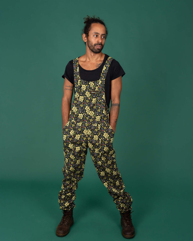 Model standing in a relaxed pose, wearing Run & Fly’s serpent garden dungarees. The yellow snake print entwined with colourful flowers and leaves creates a bold contrast against the black background. Paired with a black top and brown boots, the look is casual and eco-conscious. Ethical, UK-based small business known for quirky, sustainable designs in slow fashion.