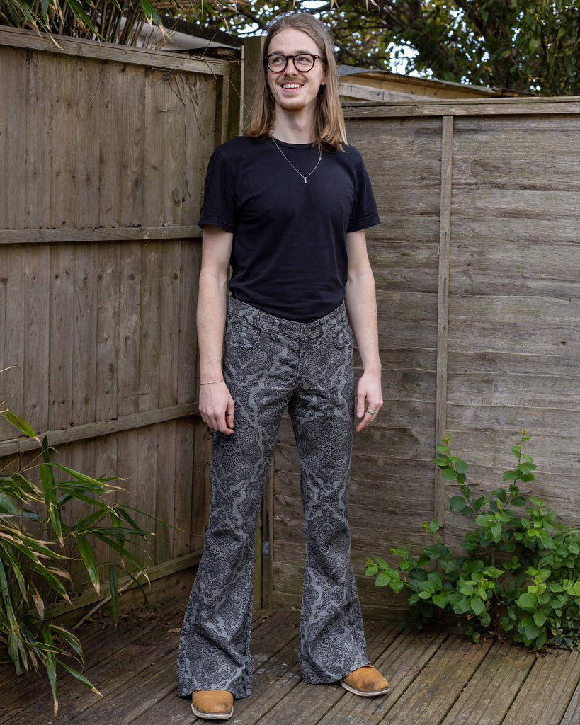  Jack stands outdoors, wearing Run & Fly’s Silver Hendrix flares with a black paisley design. He pairs the flares with a black T-shirt, smiling and looking relaxed against a wooden fence and greenery.