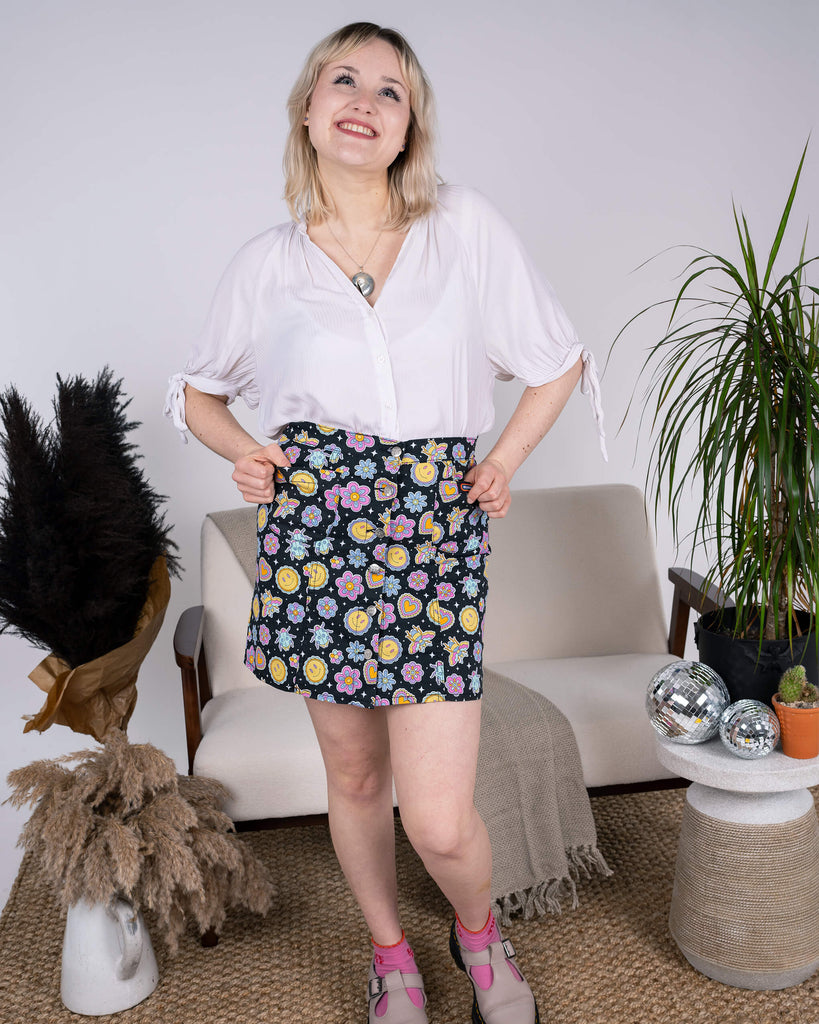  The model smiles brightly while wearing the Bee Happy A-Line Skirt by Run & Fly, paired with pink socks and platform shoes. The playful print of bees, flowers, and smiley suns creates a vibrant look. Ethical slow fashion that’s fun and full of joy.