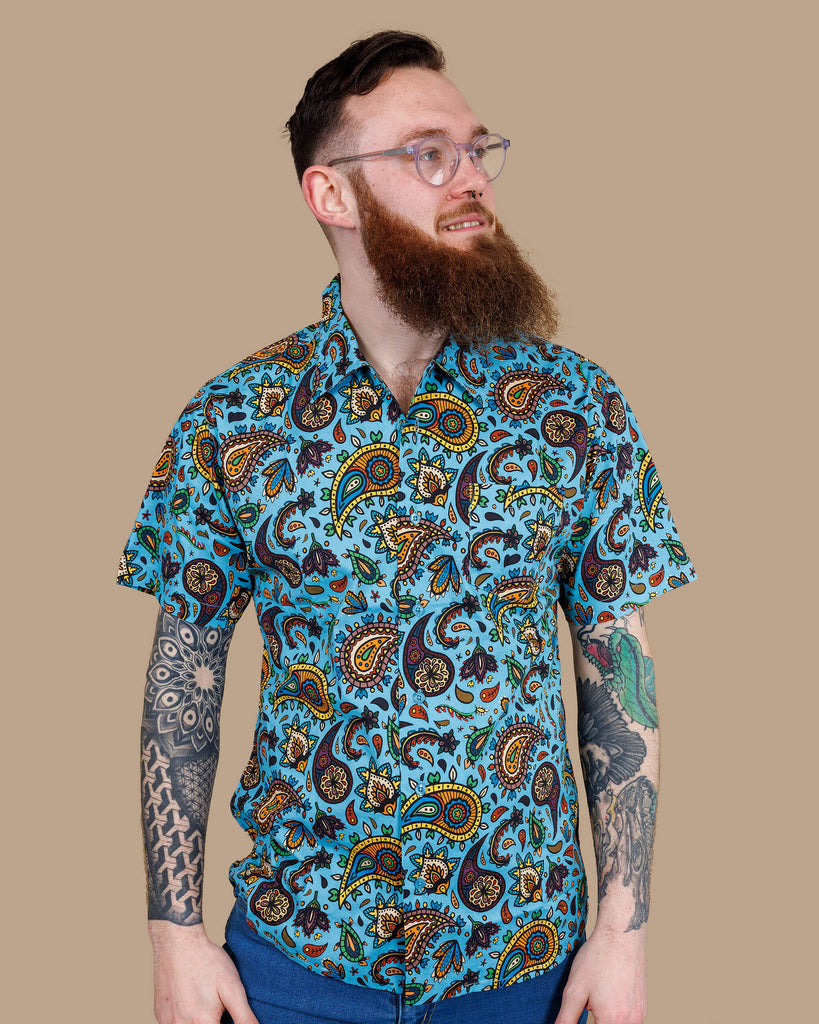 Front view of a bearded model in the Run & Fly Sky Blue Paisley Short Sleeve Shirt. The shirt features intricate paisley designs in earthy colours on a blue background. Made ethically by Run & Fly, a slow fashion brand.