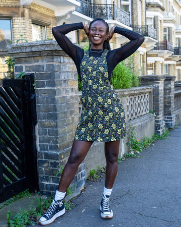 A model is stood outside in Hove wearing Run & Fly Halloween: Serpent Garden Stretch Twill Pinafore Dress with a long sleeved black mesh top underneath. The dark base of the pinafore is covered with vibrant yellow snake designs, surrounded by small floral patterns, creating a quirky, eye-catching look. The model pairs the outfit with black high-top trainers and is smiling at the camera with her hands in her hair