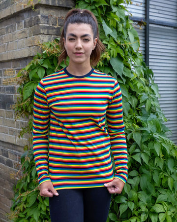 Sophie is wearing Retro Rainbow Brights Repeat Striped Long Sleeved T Shirt with black trousers. She is stood outside in Hove in front of a brick wall with greenery and is posing facing the camera while holding the bottom of the t shirt.