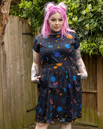 Zoe is a tattooed white female with pink long hair and is wearing a galaxy print space planets tea dress in a garden setting.