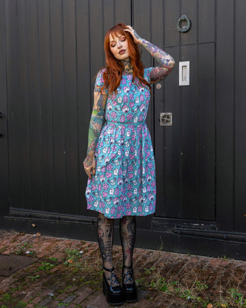 Full-length photo of a red-haired woman posing in front of a black wooden door, showcasing her colourful tattoos and wearing a playful blue dress with a ghost and crystal print by Run & Fly. The brand is celebrated for its ethical and slow fashion practices.