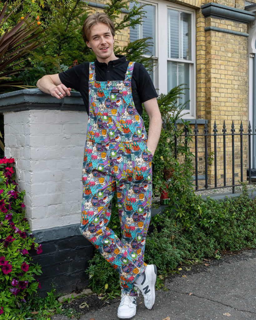 Jack is stood outside in Hove by greenery  wearing Run & Fly Halloween: Spooky Stuff Stretch Twill Dungarees with a short sleeve black top underneath. The light blue dungarees have an all over print of various Halloween themed items such as skulls, snakes, eyeballs, ghosts, bats, crows, candles, potions, crystals and more. Jack is smiling facing the camera with his hands in his pockets. 