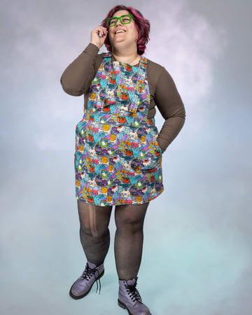 Jackie is seen in a playful pose, wearing the Pretty Spooky Pinafore with its vibrant spooky design. The long-sleeve brown top, patterned tights, and boots complete the look, showcasing the versatile and fun aesthetic of the outfit.