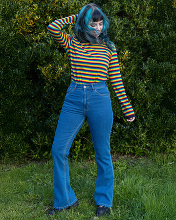 Faeryn is stood in a garden area wearing the unisex stonewashed bellbottom flares leg high waisted jeans with the Retro Rainbow Brights Repeat Striped Long Sleeved T Shirt and black trainers. They are facing forward posing with one leg bent out to show off the flares and one hand behind their head whilst looking down to the right.