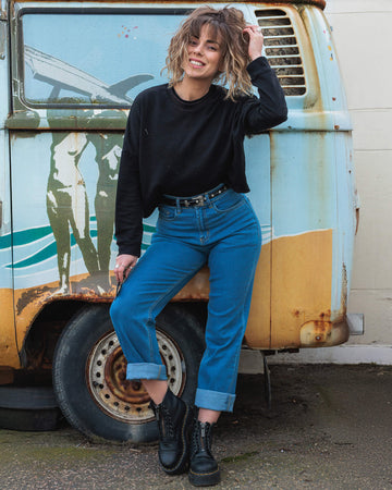  Hannah smiles while posing in front of a vintage van, wearing Run & Fly’s blue straight-leg jeans with a black sweatshirt and star-studded belt. Her look is completed with black chunky boots, giving off a relaxed, cool vibe.