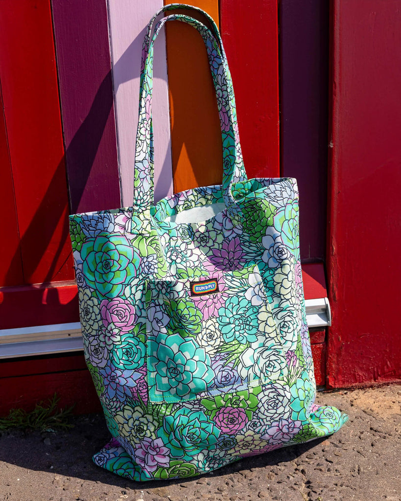 The Succulents Tote Bag leaning up against a colourful beach hut along hove seafront. The tote bag has a front pocket with a rainbow run and fly label in the top middle. The succulent print features pastel blue, pastel teal, green, purple and pale yellow toned succulent plants with black outlines.