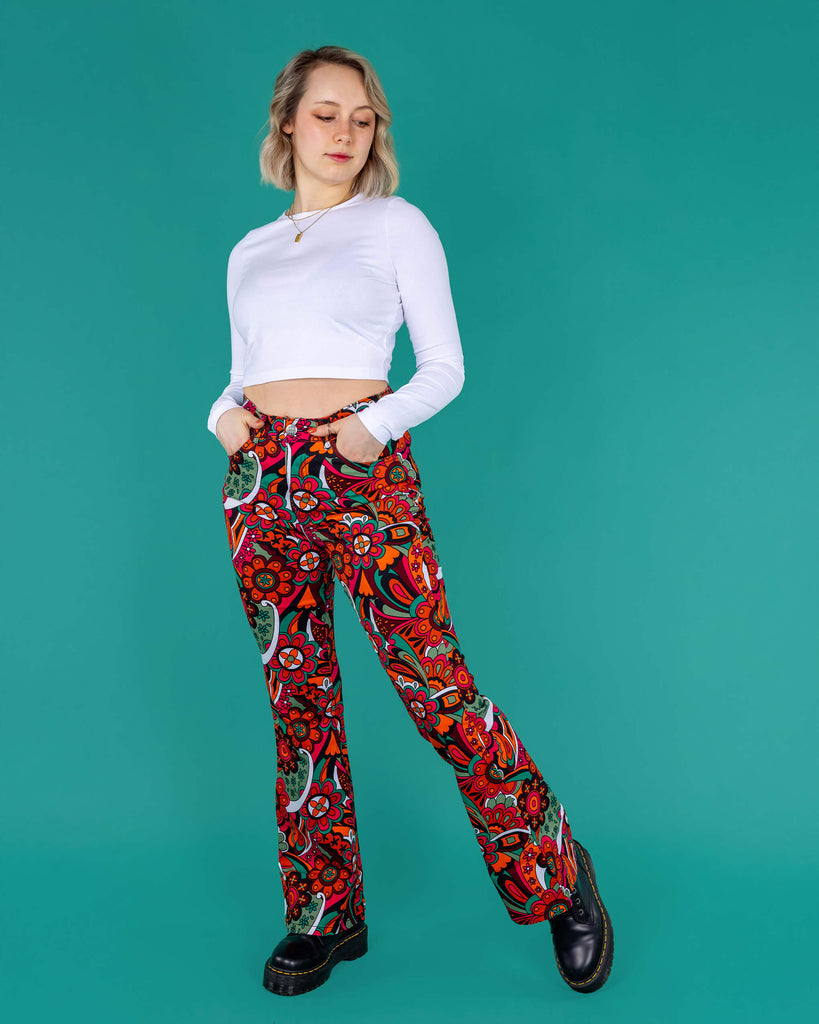 Amy a blonde model with bobbed hair is wearing a white long sleeved cropped tee with jazzy 70's swirly flares with black boots on a green background 