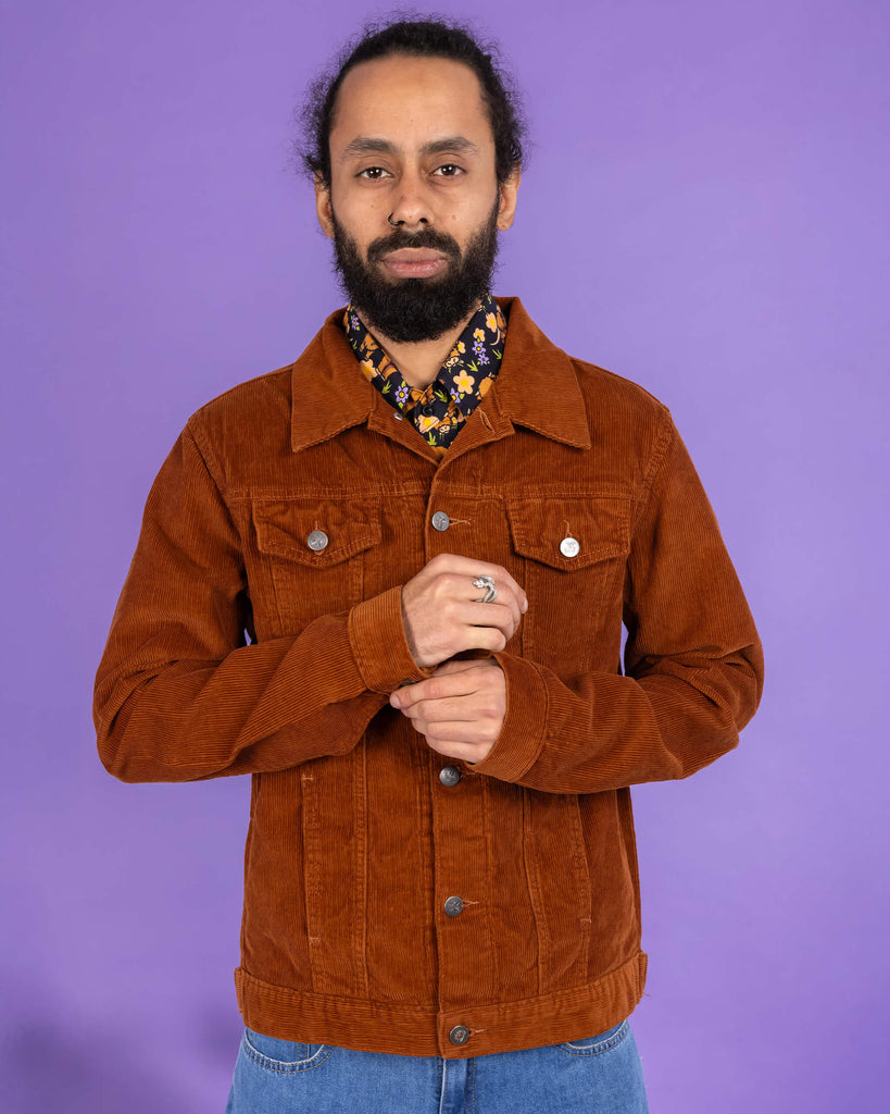 Richard, a hispanic male model with dark hair in a bun and a beard, is stood in a photography studio in Hove in front of a lilac backdrop wearing Retro Vintage Tan Corduroy Unisex Western Jacket buttoned up with Highland Cows Short Sleeve Shirt underneath and blue jeans. Richard is posing to the camera and is doing up the button one of the jacket sleeves. The photo is cropped at the thighs.